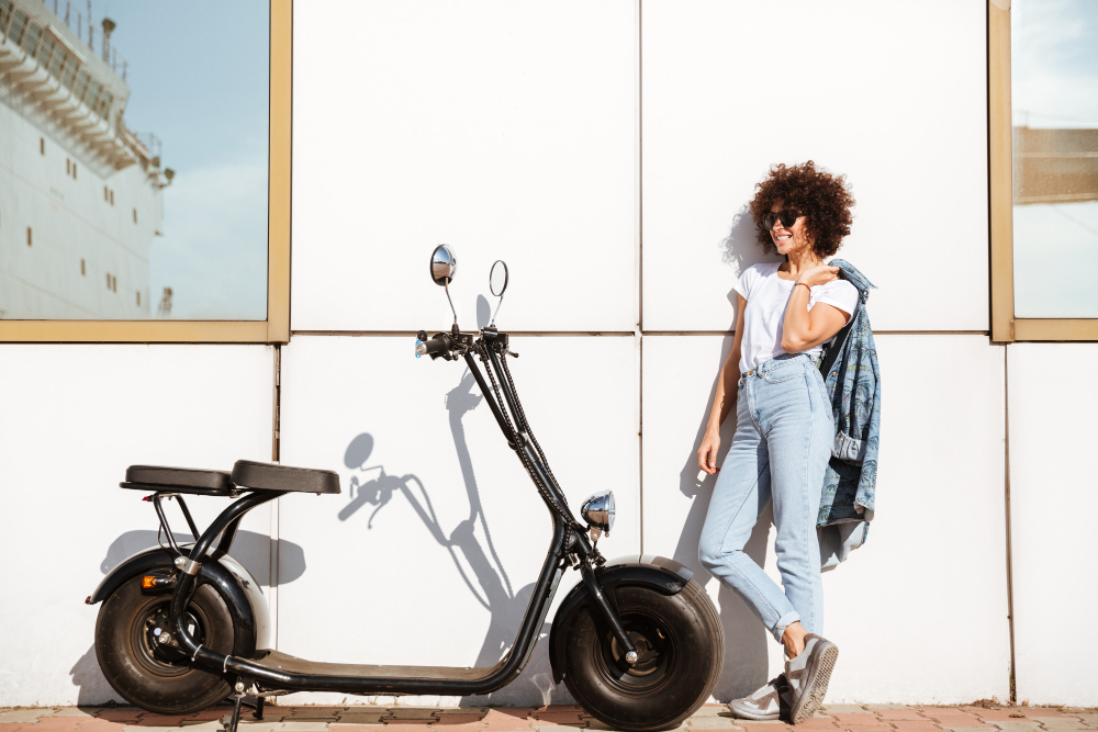 Mulher sorridente ao lado de uma scooter elétrica estacionada em frente a uma parede branca, representando mobilidade urbana sustentável. 