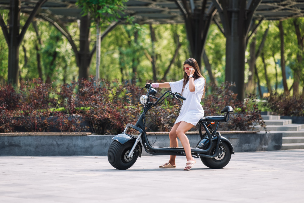 Mulher pilotando scooter elétrica em área urbana arborizada, usando vestido branco e óculos de sol. 