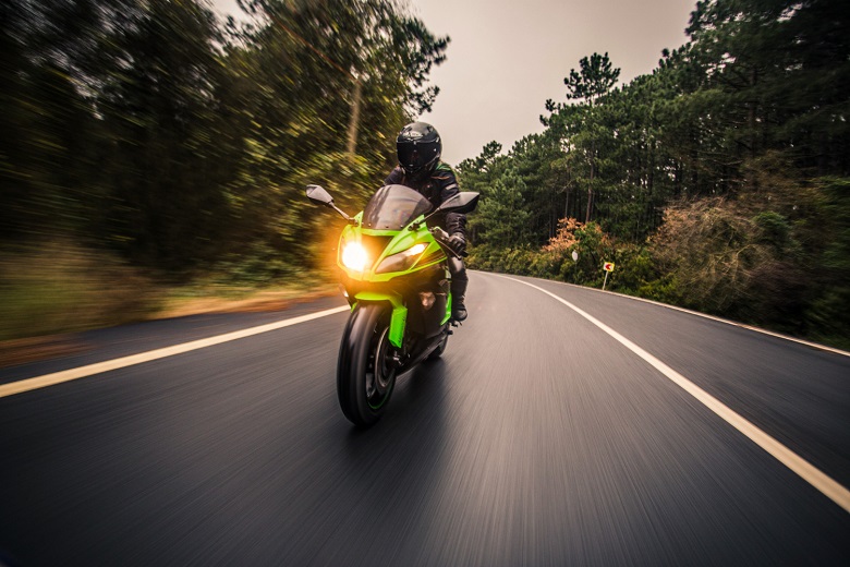 “Motociclista pilotando uma moto esportiva verde em alta velocidade por uma estrada cercada por árvores, ilustrando o tema ‘moto elétrica ou flex: qual é a melhor opção?’, que compara desempenho, economia e sustentabilidade entre diferentes tipos de motocicletas.”