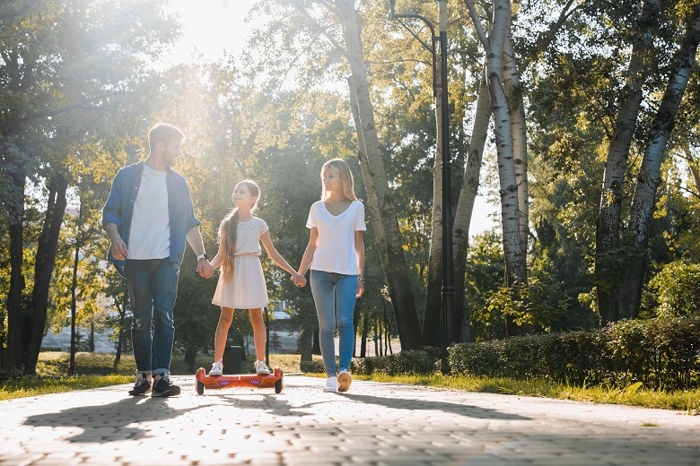 Little smiling girl learning to ride a hoverboard with her parents outdoors.