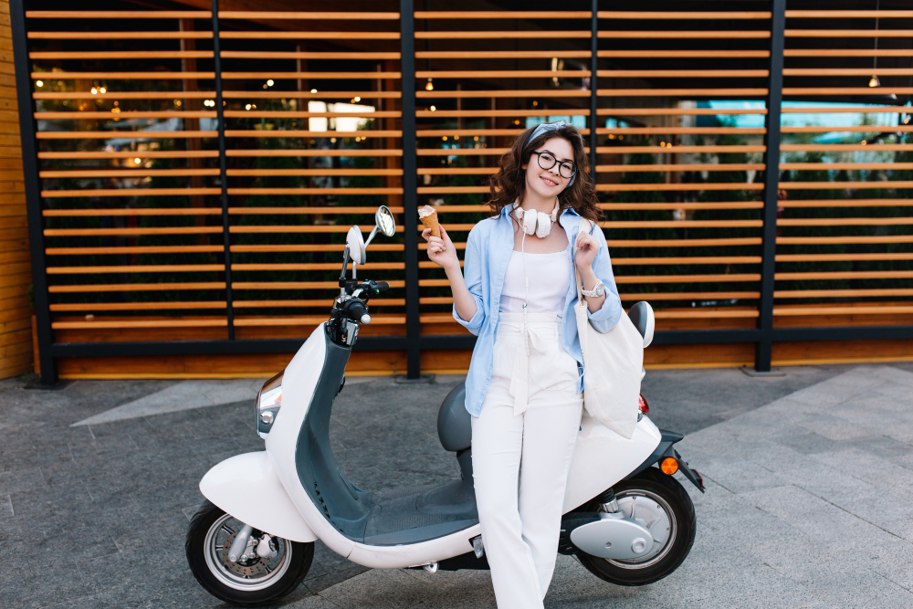 “Mulher jovem sorridente em pé ao lado de uma scooter branca, simbolizando mobilidade urbana sustentável. A cena ilustra o tema ‘qual a velocidade de uma moto elétrica’, destacando estilo, praticidade e modernidade no uso desse tipo de veículo.”